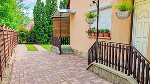 Un chemin pavé mène à un bâtiment entouré d’une clôture en bois, agrémenté de plantes en pot et doté d’un espace abrité avec un banc.