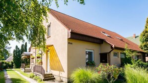 Une maison à deux étages, avec un toit recouvert de tuiles rouges, une porte jaune et un petit porche agrémenté d’un banc.