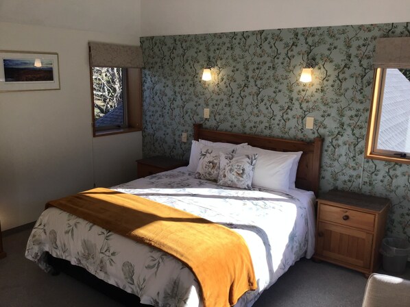 Quarto com cama, duas mesas de cabeceira, cabeceira de madeira, papel de parede floral, duas luzes fixadas na parede, uma janela e um quadro na parede.