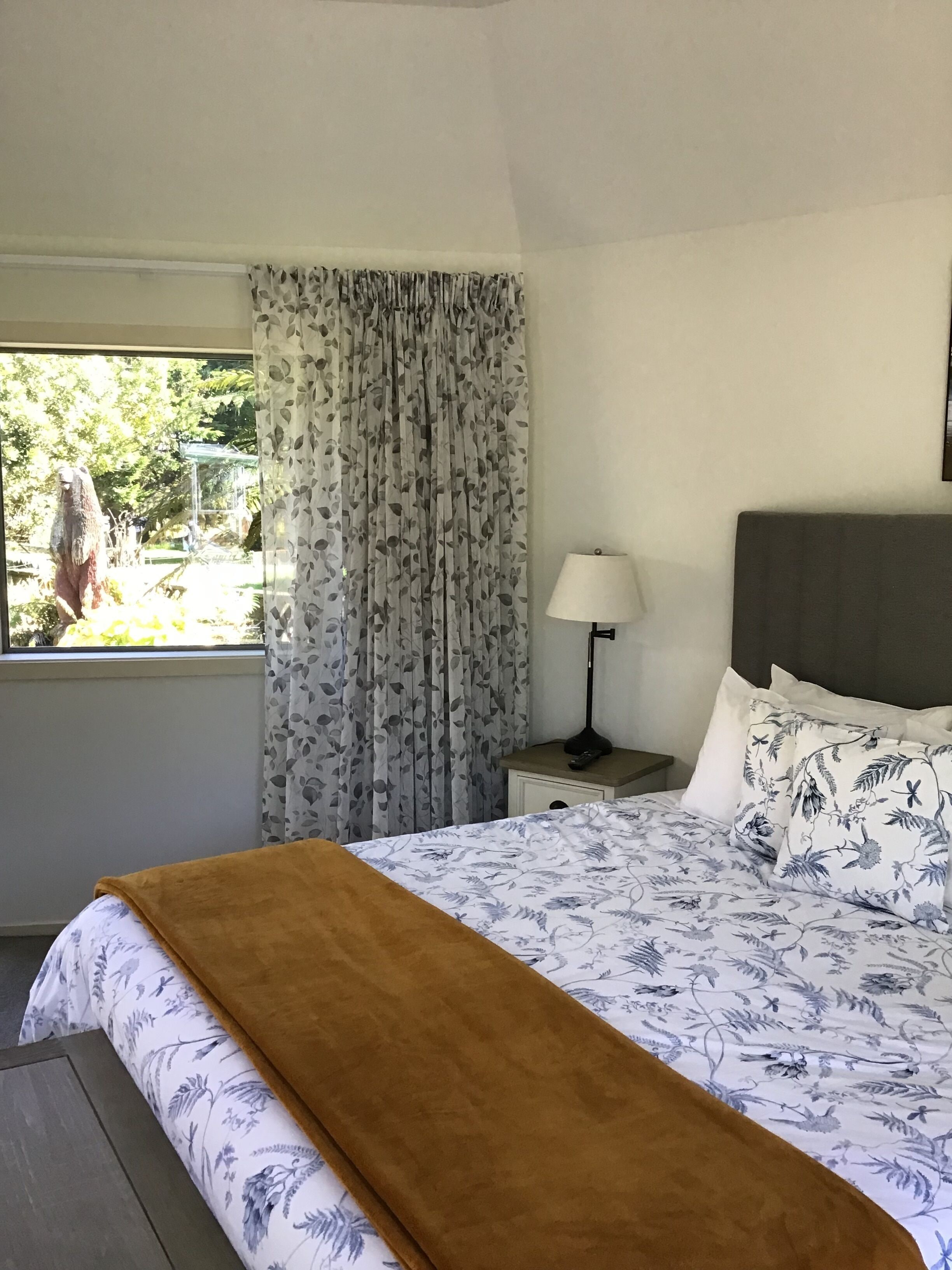 A bedroom with a bed, a nightstand, a lamp, a window with curtains, and a view of greenery outside.