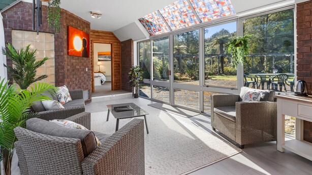 A modern outdoor lounge area with wicker furniture, a glass door leading to a garden, and a brick wall with a fireplace. - Woodland Glen Lodge (Hokitika)