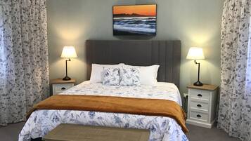 Um quarto com uma cama, duas mesinhas de cabeceira, um criado-mudo, um banquinho e um quadro com um pôr do sol sobre o oceano.