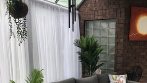 A modern living room with a grey sofa, two armchairs, a coffee table, and decorative pillows. There are potted plants, a hanging plant, and a large window with sheer curtains. - Woodland Glen Lodge (Hokitika)