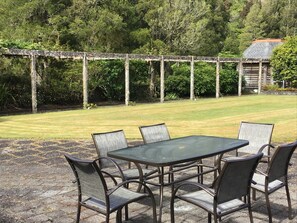 Um pátio externo com mesa e cadeiras, rodeado por uma pérgola de madeira e vegetação exuberante.