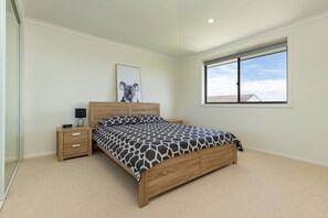 Un dormitorio con una cama, una mesita de noche, un reloj de pared, un cuadro enmarcado y una ventana con vistas al cielo y edificios.