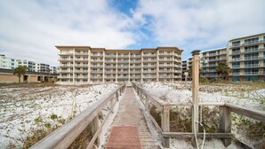 Une promenade en bois mène à un hôtel en bord de mer, doté de nombreux balcons, avec une plage de sable en arrière-plan.