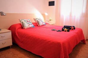 A neatly made bed with a red bedspread, two floral pillows, and a starfish decoration. There is a bedside lamp, a small table with a plant, and a window with curtains.