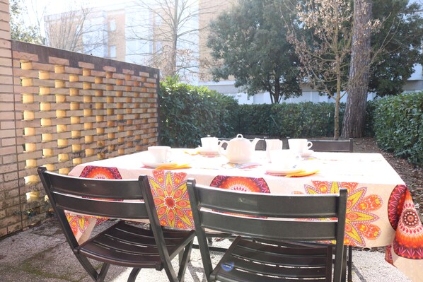 An outdoor dining area with a brick wall, a table set with white plates, cups, and a teapot, and two chairs with patterned tablecloths.