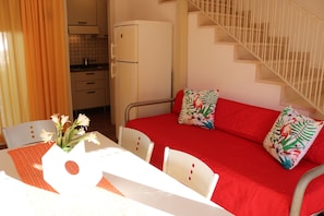 A room with a red sofa, white chairs, a small table with a vase of flowers, a kitchen area, and a staircase.