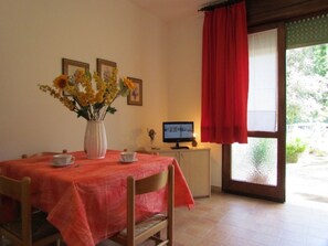 Un comedor con una mesa dispuesta para dos personas, un jarrón de flores, un televisor y una ventana con cortina roja.