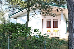 A white building with a green roof, two windows with wooden shutters, and a table with chairs outside.