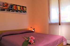 Une chambre à coucher comprenant un lit, une table de chevet, une lampe, une fenêtre avec des rideaux et une œuvre d’art murale.