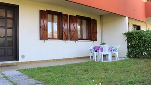 Un bâtiment à la façade blanche, avec une porte noire et un ensemble de volets en bois. On y trouve un petit jardin avec une table et des chaises.