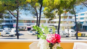A balcony with a white chair, a small table with a vase of flowers, and a view of a street with parked cars and buildings. - Spacious Apartment With Large Balcony (San Michele al Tagliamento)
