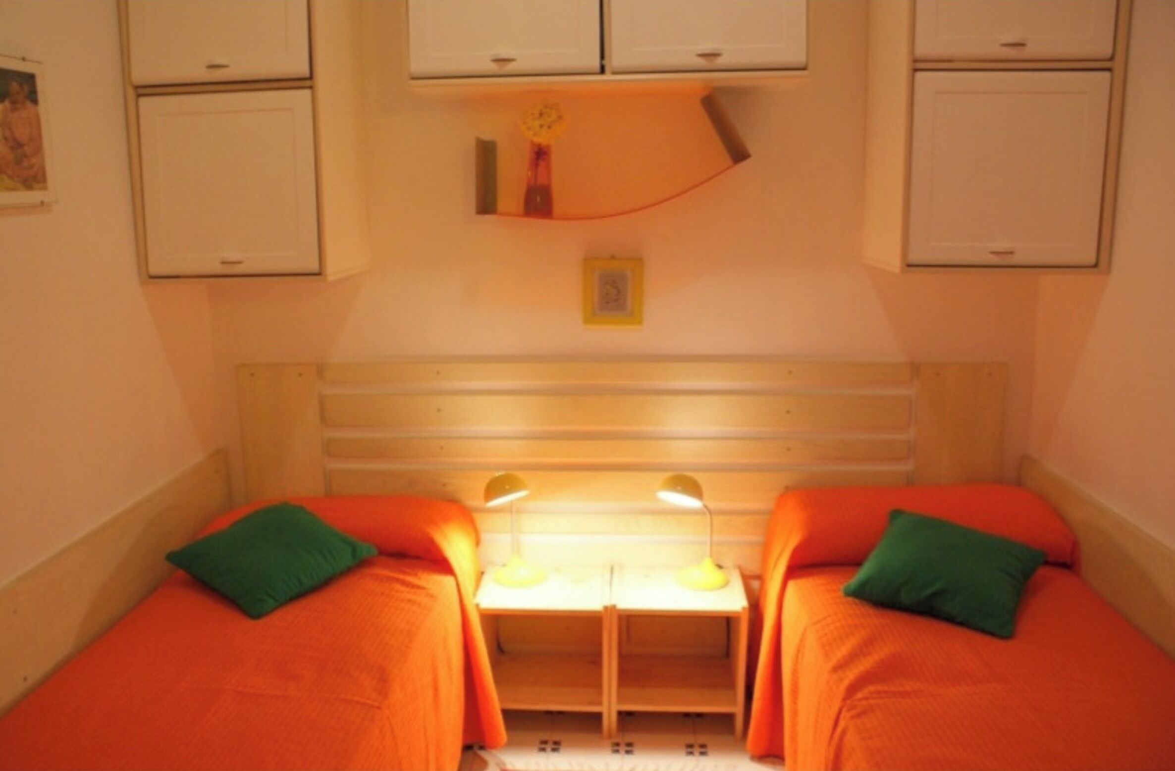 Une petite pièce, de taille modeste, avec deux lits, chacun recouvert d’une couverture orange et agrémenté d’un oreiller vert. Au centre, on trouve un petit bureau avec deux lampes, et une étagère au-dessus du bureau, sur laquelle sont posés quelques objets.