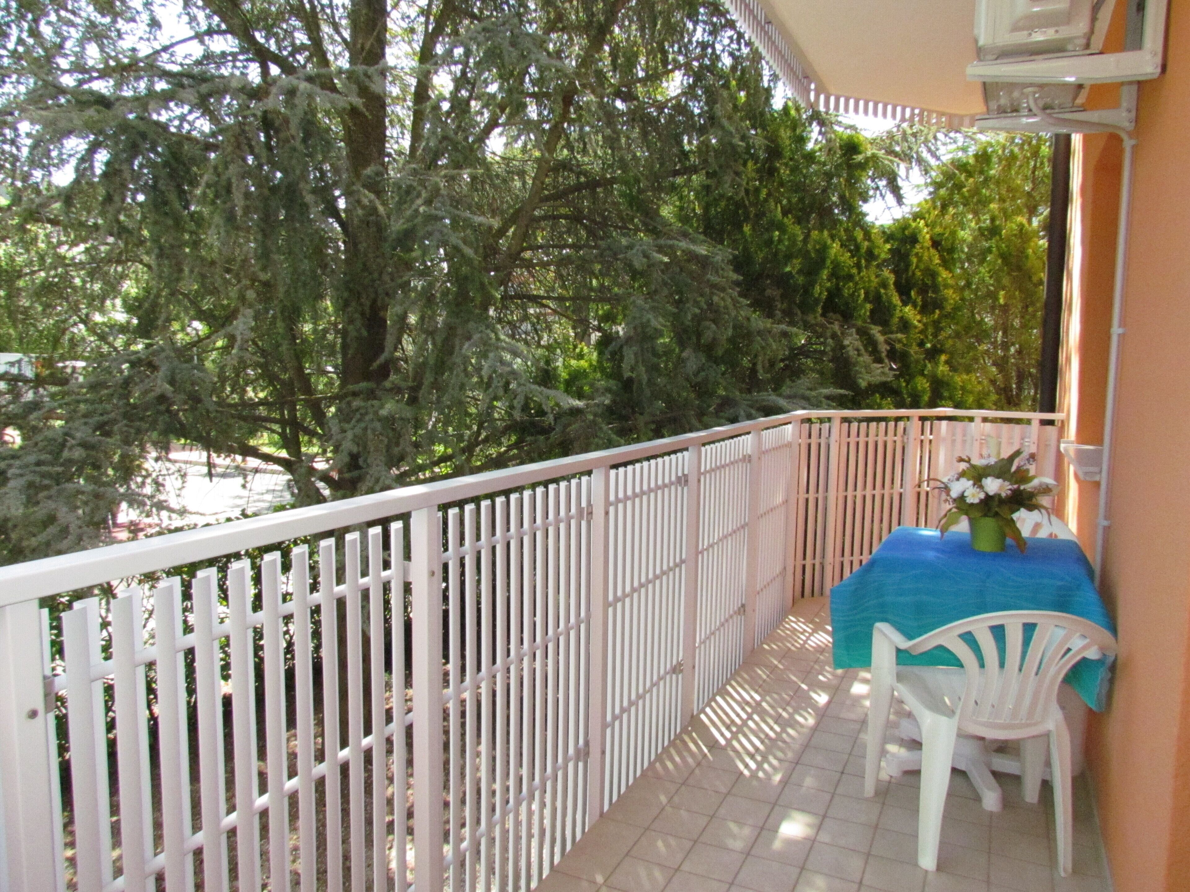 Un balcon avec une balustrade blanche, une petite table recouverte d’une nappe bleue et une chaise blanche.