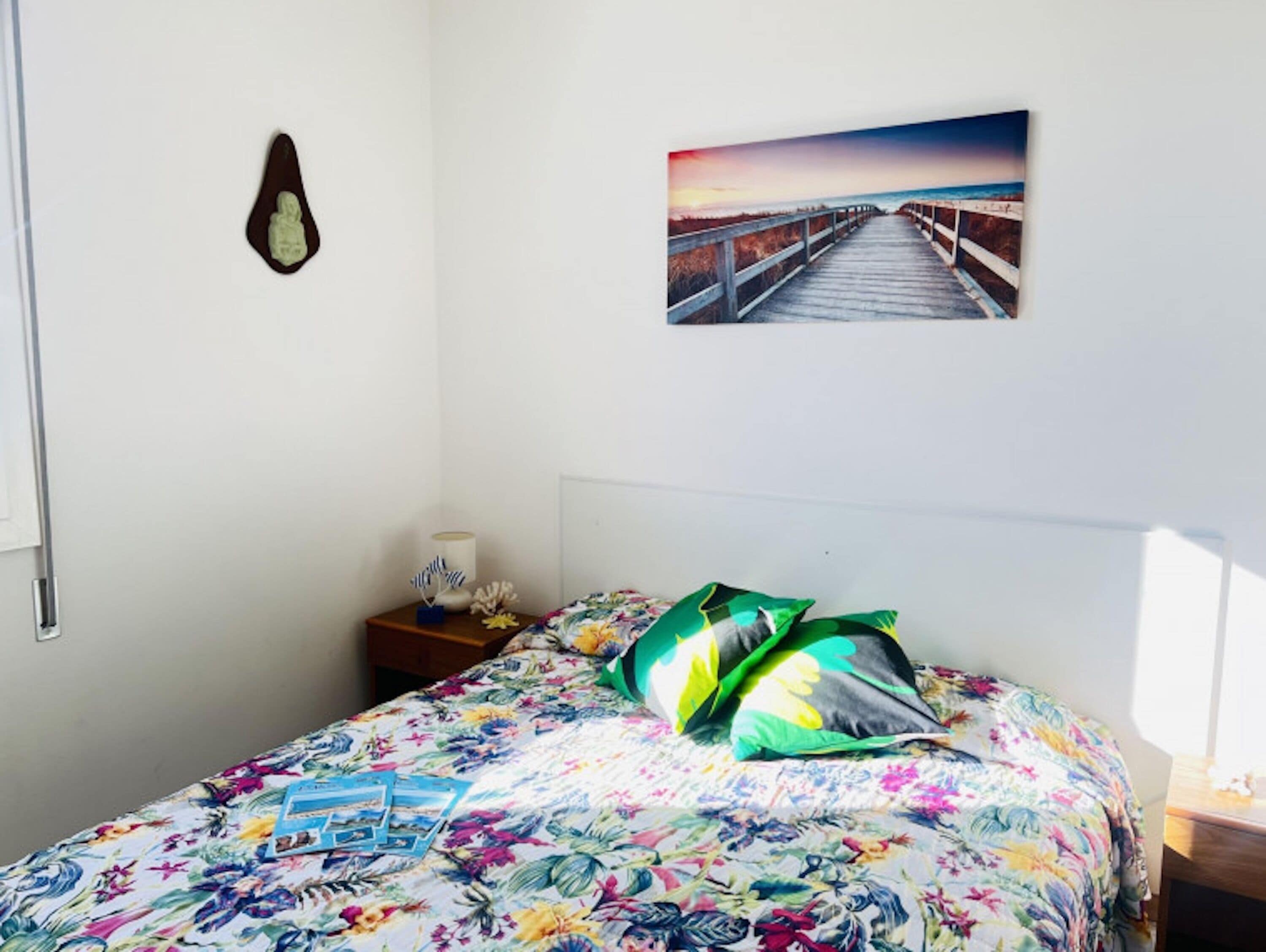 Een slaapkamer met een bloemenbedekker, een schilderij van een pier, een klein nachtkastje met een lamp en een wandklok.