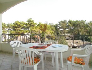 Un balcon avec une table ronde et des chaises blanches, donnant sur un quartier résidentiel bordé d’arbres.