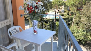 A balcony with a white table and chairs, a vase of flowers, and a view of a pool and greenery. - Stay Just few Meters From the Beach (San Michele al Tagliamento)