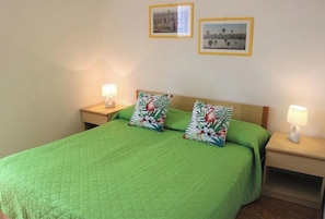 Ein Einzelbett mit einer grünen Steude, zwei floralen Kissen und zwei Nachttischlampen auf Holzbeistelltischen.
