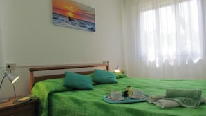 Ein Schlafzimmer mit grüner Bettdecke, einem Nachttisch mit Lampe, einem Sonnenuntergangsbild und einem Fenster mit durchsichtigen Vorhängen.