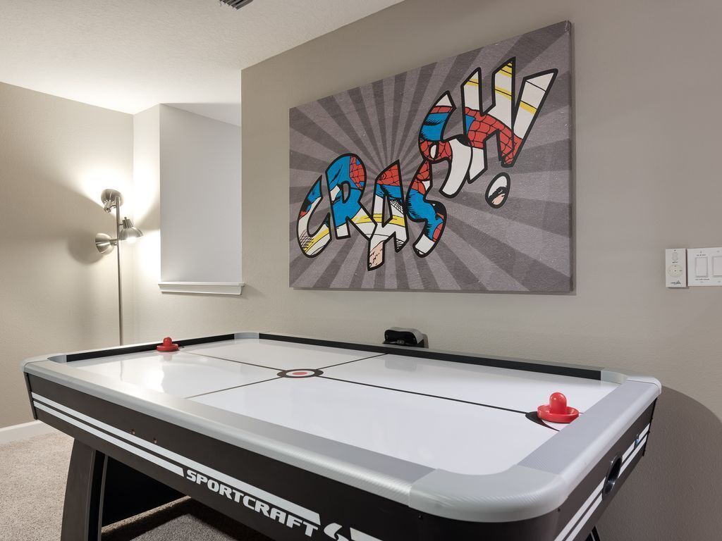 A pool table with a 'SPORTCRAFT' logo, a wall-mounted artwork with the word 'CRASH!' in a comic style, and a floor lamp.