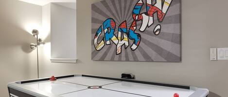 A pool table with a 'SPORTCRAFT' logo, a wall-mounted artwork with the word 'CRASH!' in a comic style, and a floor lamp.