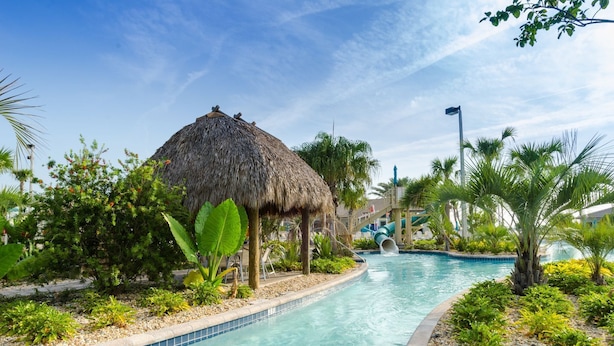 Une piscine dotée d’une structure surmontée d’un toit de chaume, entourée de plantes tropicales et agrémentée d’un toboggan aquatique.