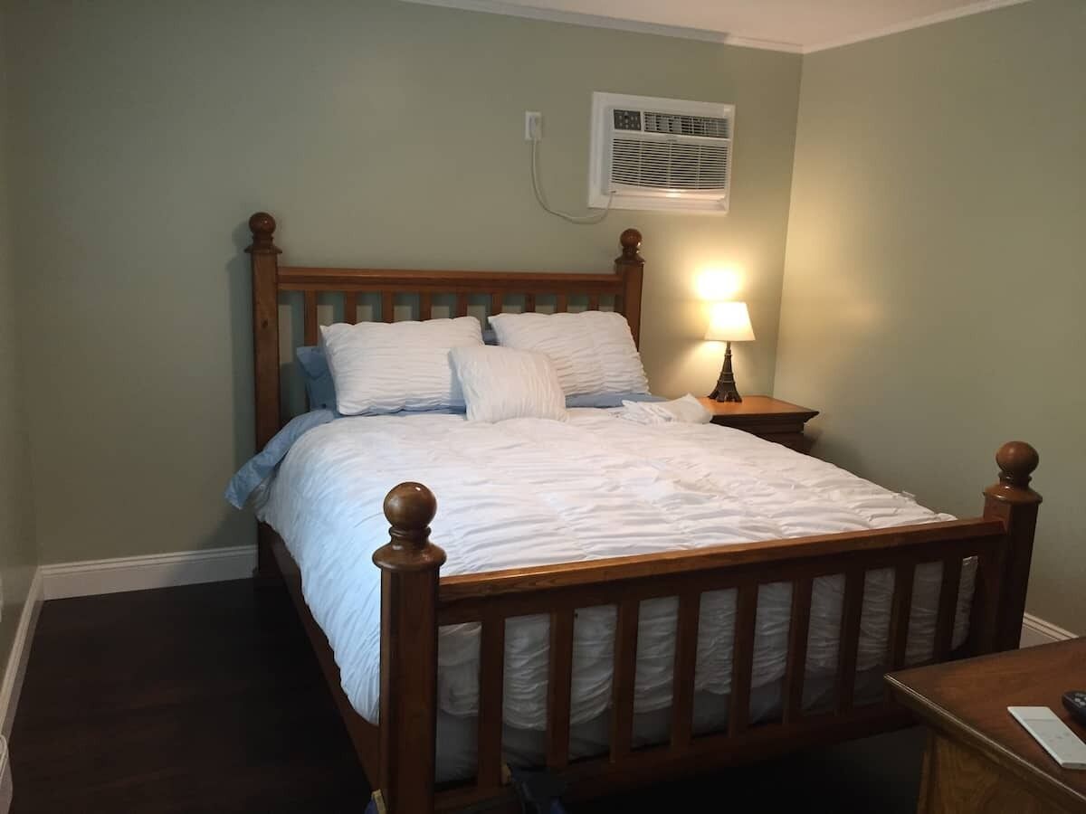 A single bed with a wooden frame and white bedding, a bedside table with a lamp, and a wall-mounted air conditioning unit.