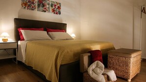 A bedroom with a bed, bedside tables, a nightstand, a woven ottoman, and wall art. - Palazzo Nicastro Guest House Red (Avola)
