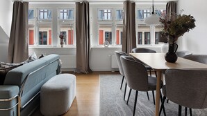 A modern living room with a sofa, dining table, and chairs, large windows with curtains, and a vase with flowers on the windowsill.