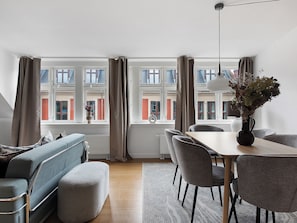Ein modernes Wohnzimmer mit einer Couch, einem Esstisch und Stühlen, großen Fenstern mit Vorhängen und einer Vase mit Blumen auf der Fensterbank.