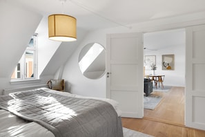 Quarto com cama, espelho redondo, luminária no teto e vista para uma sala de jantar.