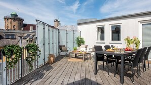 A rooftop terrace with a dining table and chairs, surrounded by a glass enclosure, and a view of a historic building in the background.