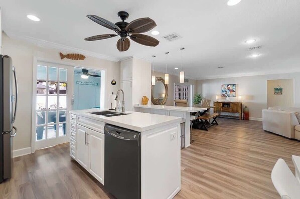 A modern kitchen with white cabinetry, a dishwasher, a ceiling fan, and a dining area with a table and chairs. - Tiki Retreat! Pool, large game room, 5min to FLL and BEACH! (Dania Beach)