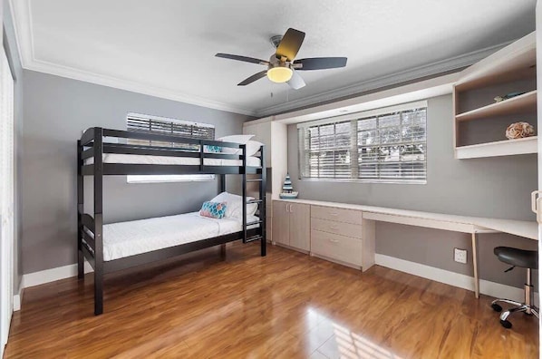 Un lit superposé avec un bureau et une chaise, un ventilateur de plafond et une fenêtre avec des stores.