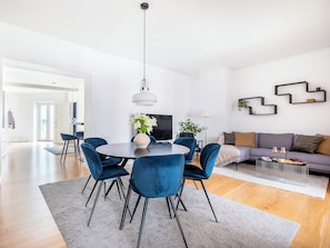 Uma sala de estar moderna com uma mesa de jantar redonda, cadeiras de veludo azul, uma TV de tela plana, um sofá e um objeto decorativo na parede.