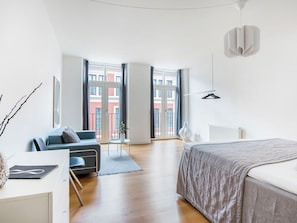 Quarto moderno com cama, sofá, radiador e vista para edifícios através de grandes janelas.