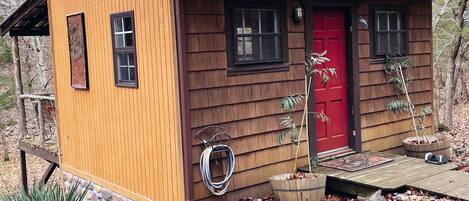 A small wooden cabin with a red door, two windows, and a porch area.