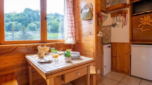 A wooden cabin kitchen with a dining table set for breakfast, a window with a view of greenery, and a refrigerator. - Soyouz Vanguard by Interhome (Le Corbier)