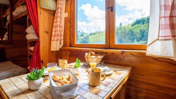 A cozy cabin interior with a wooden interior, a window with a view of greenery, a table with breakfast items, and a basket of bread. - Soyouz Vanguard by Interhome (Le Corbier)