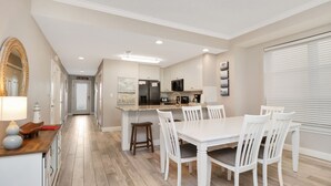 A modern kitchen with white cabinetry, a central island, and a dining area with white chairs. - Great Views, Rooftop Pool, Hot Tub & Sauna ~ Summerhouse 106 (Mexico Beach)