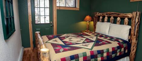 Een slaapkamer met een houten hoofdbord, een gewatteerde sprei, een nachtkastje met een lamp, en een raam met jaloezieën.