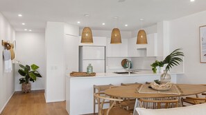Una cocina moderna con gabinetes blancos, una mesa de comedor de madera, lámparas colgantes de mimbre y una planta en maceta.