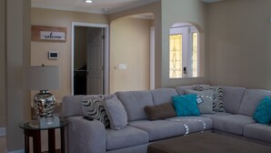 En stue med en grå sofa, et lille sofabord og en lampe. Der er et skilt med ordet 'welcome' på væggen, og et vindue med dekorativt glas.