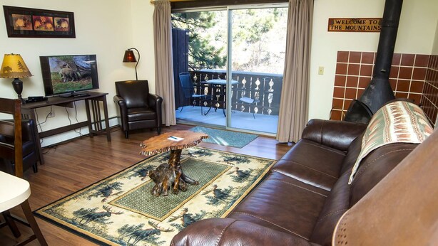 Un salon chaleureux avec un canapé en cuir marron, une table basse en bois et un téléviseur posé sur un meuble en bois. Une grande fenêtre offre une vue sur les arbres, et une pancarte indique : « Bienvenue à la montagne ».