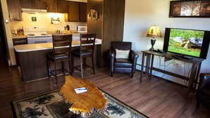 Une cuisine moderne avec des meubles de couleur bois foncé, un îlot central doté de tabourets de bar, un coin repas avec un fauteuil en cuir, un téléviseur posé sur un meuble en bois et un tapis orné de motifs naturels.