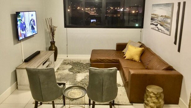 A modern living room with a brown leather sofa, two grey chairs, a small round table, a TV on the wall, and a large window with a city view. - Secure, Modern & Comfy, Lux Suite @77 Independence (Windhoek)