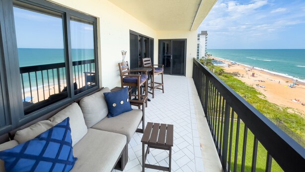 Varanda com sofá, cadeiras e mesa, com vista para uma praia e uma visão clara do oceano.