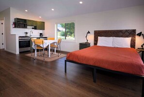 A bedroom with a bed, a small kitchenette, a dining area with a table and chairs, and a window providing a view of greenery.